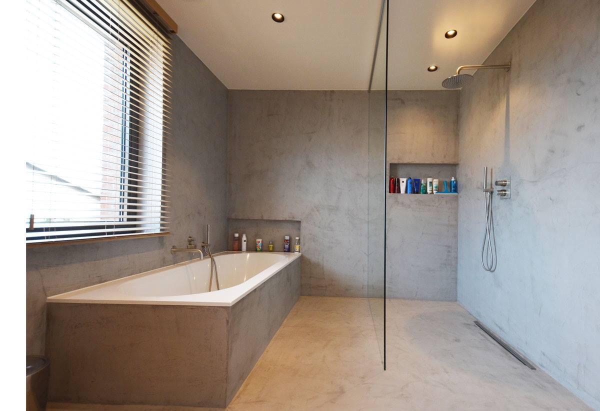 Moderne badkamer met grijze naadloze wanden, een ingebouwd wit bad onder raam met jaloezieën en een inloopdouche met glazen wand.